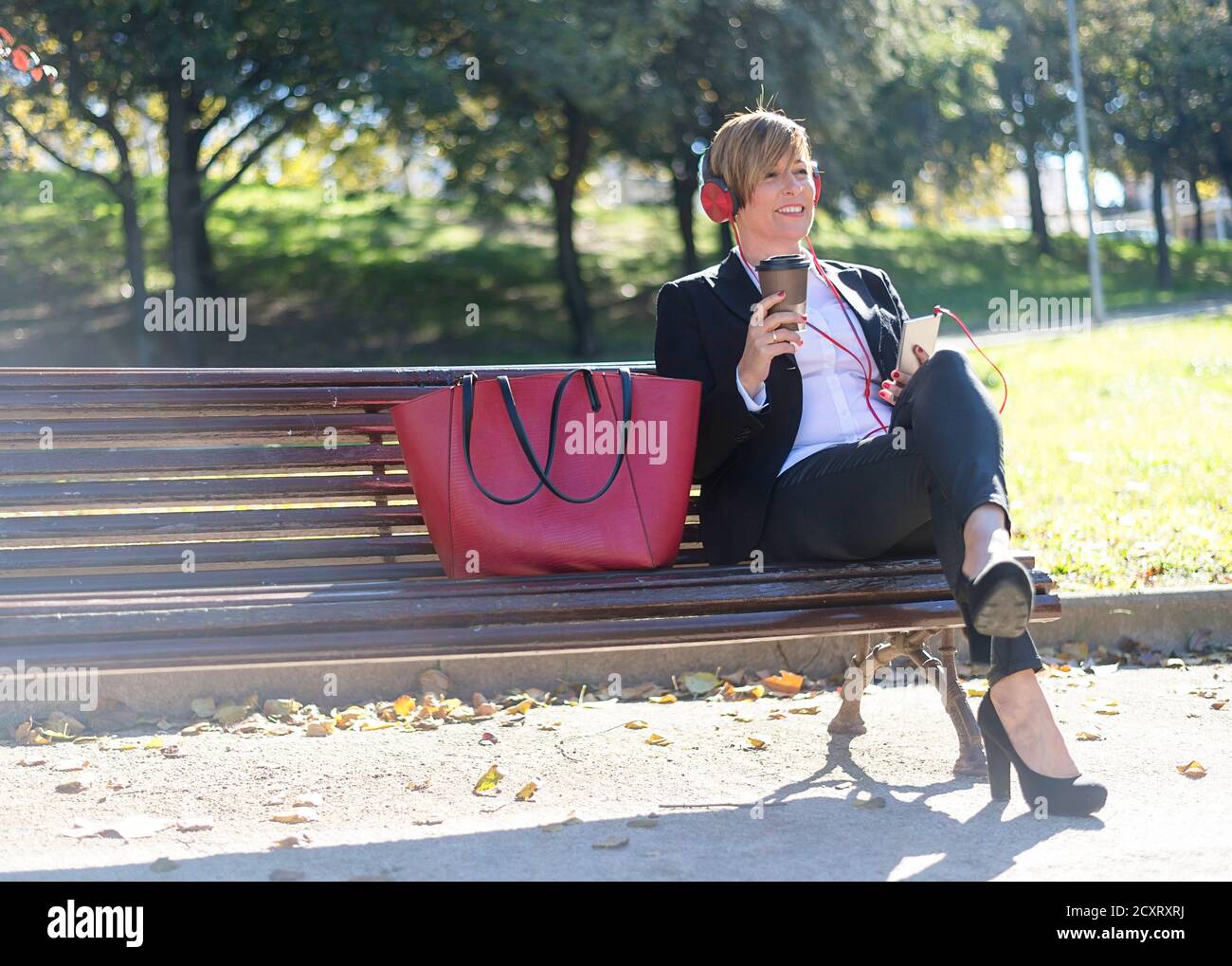 Smiling business woman sitting on a park bench while listening music by headphones and holding a take away coffee Stock Photo