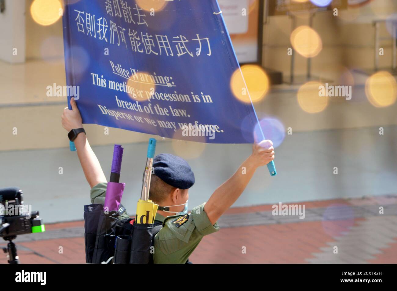 A Hong Kong Police officer holds a warning banner in Causeway Bay on National Day, 1 October 2020. Bokeh caused by mall decorations in foreground. Stock Photo
