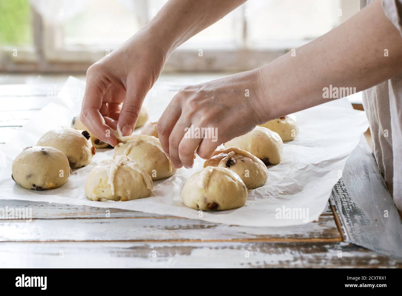 Raw unbaked buns. Ready to bake homemade Easter traditional hot cross buns on baking paper over white wooden table. Window at background. Female hands Stock Photo