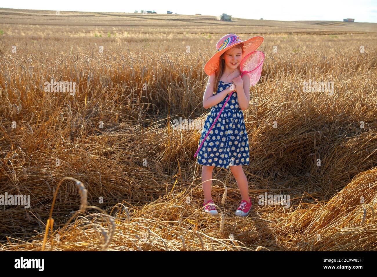 A little girl in a pink hat stands in a field and holds a net for ...