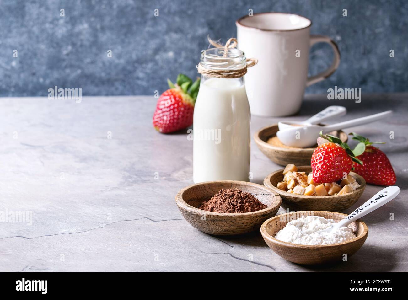 Ingredients for cooking chocolate mug cakes. Flour, cocoa powder, sugar, caramel in wooden bowls, milk, strawberries and mint served with spoons and m Stock Photo