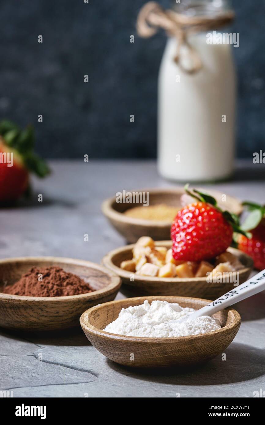 Ingredients for cooking chocolate mug cakes. Flour, cocoa powder, sugar, caramel in wooden bowls, milk, strawberries and mint served with spoons and m Stock Photo