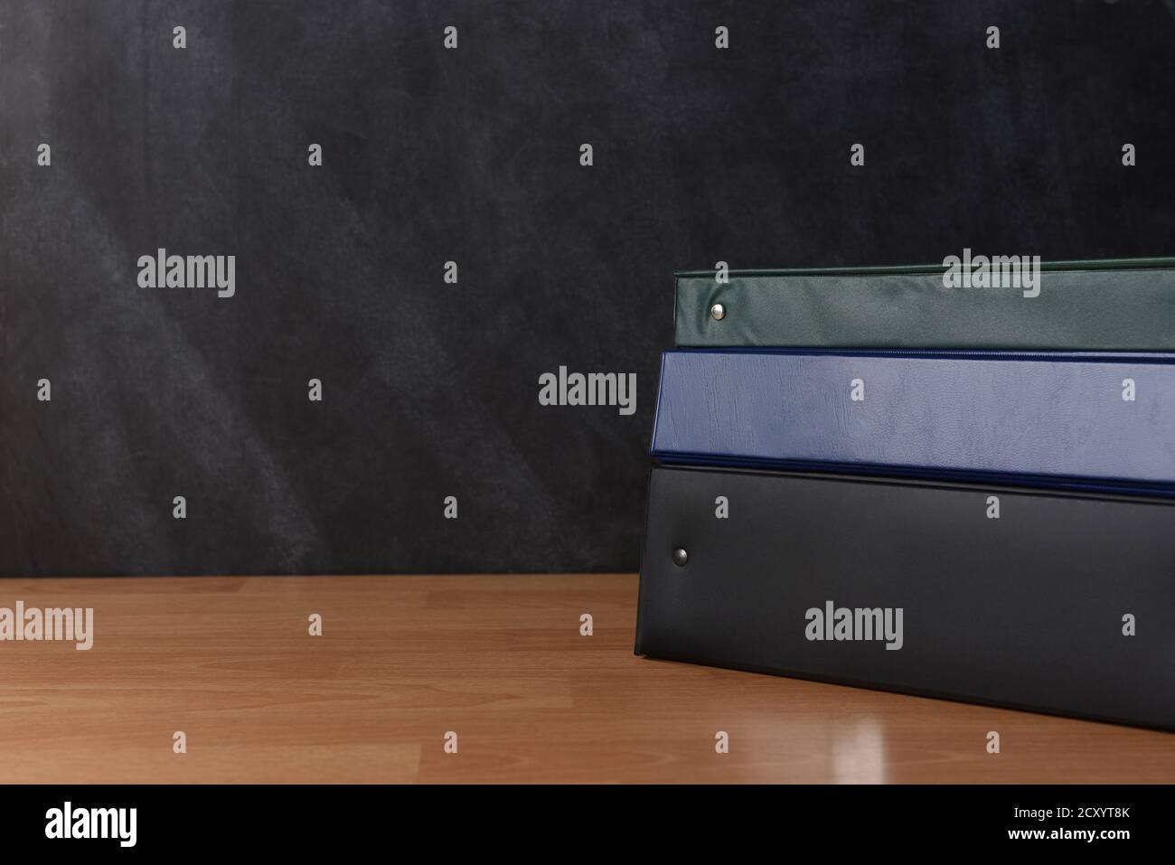 A stack of different colored binders on a teachers desk in front of a chalkboard. Stock Photo