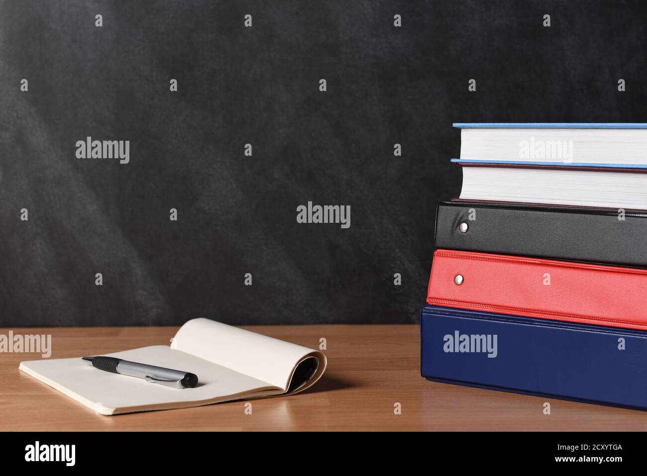 A stack of three different binders on desk in front of black board with two books and note pad and pen. Stock Photo