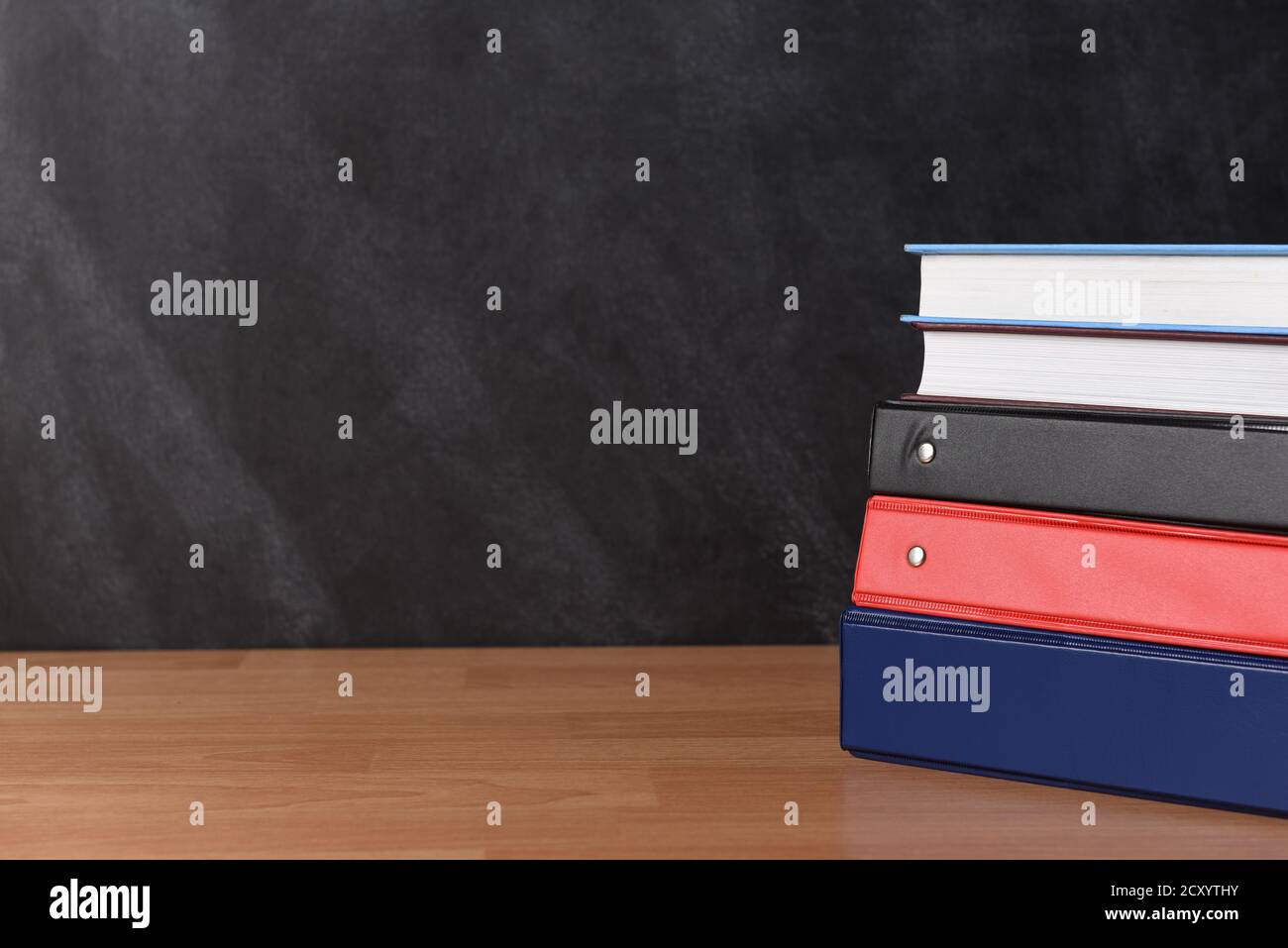 A stack of three different binders on desk in front of black board with two books. Stock Photo