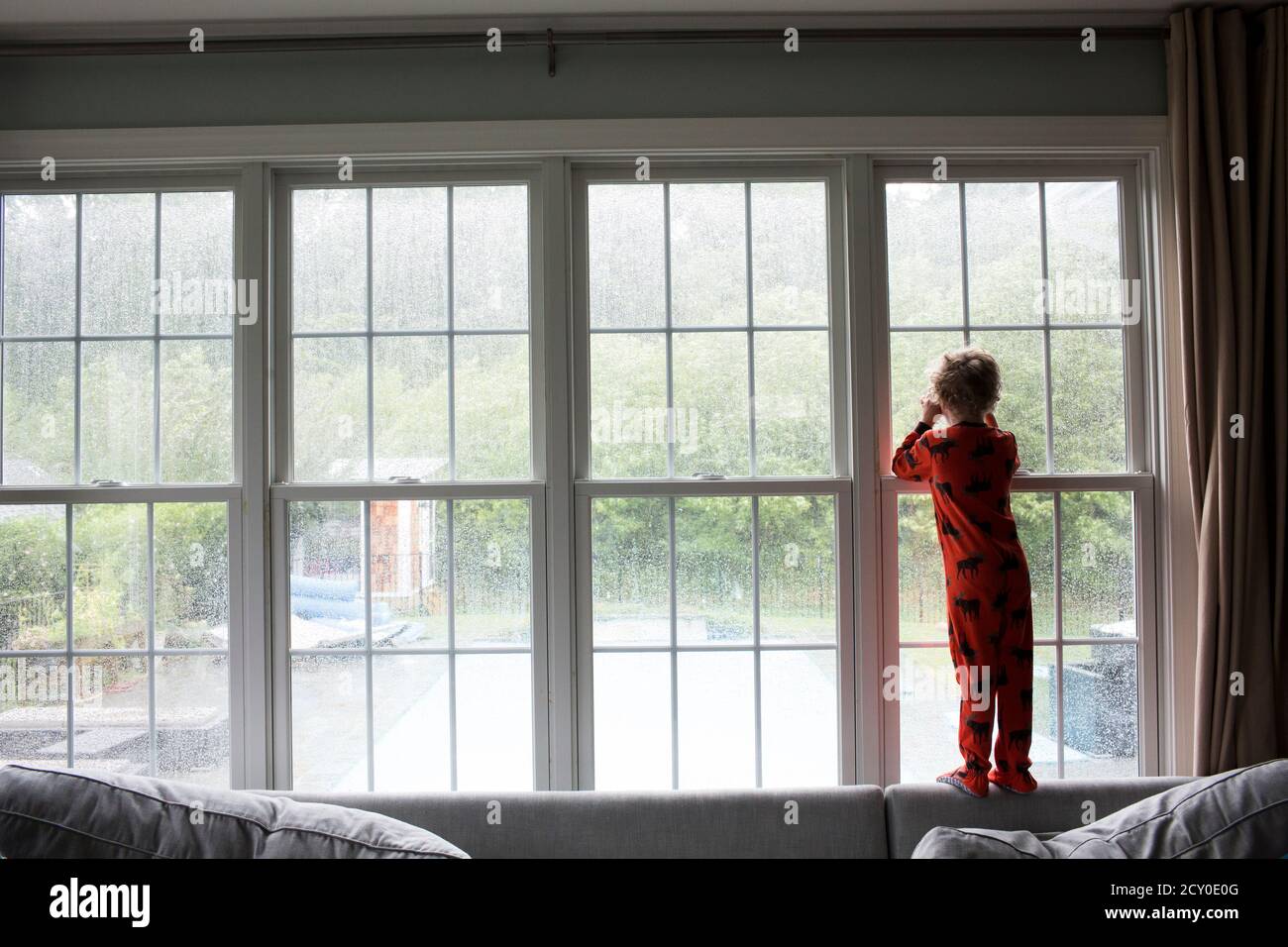 Wide Rear View of Boy in Red Pajamas Looking Out Window at Rain Stock