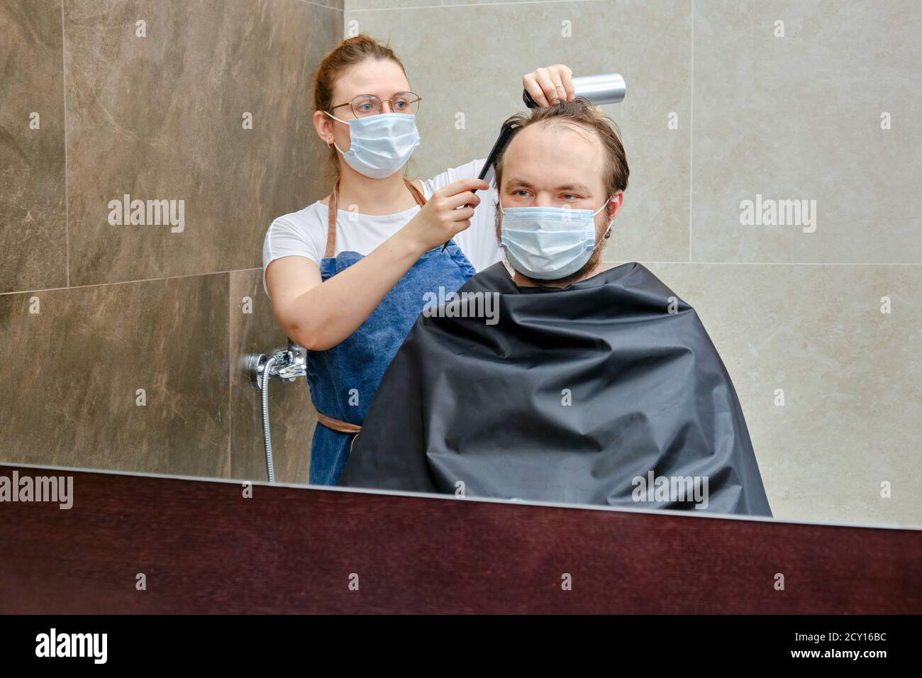Woman in medical mask creates hairstyle for man with comb and hair spray in home bathroom, copy space. Concept of closed hairdressers and barbershops Stock Photo