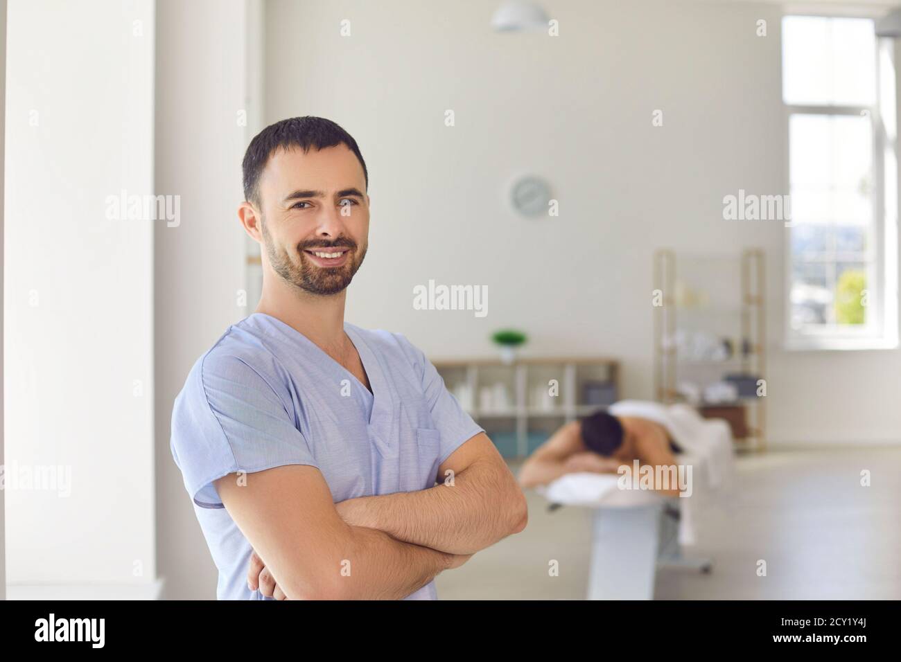 Smiling masseur or physiotherapist standing arms folded in massage room of modern health center Stock Photo