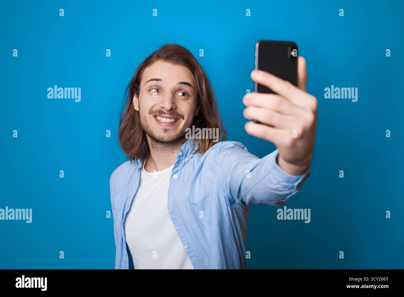 Young long haired man with beard is smiling and making a selfie using a phone on a blue studio wall in casual clothes Stock Photo