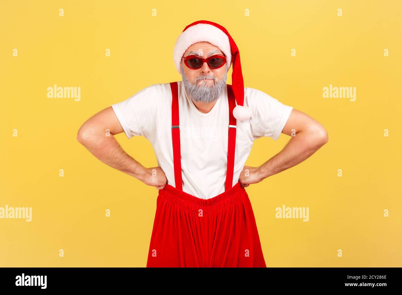 Funny adult man with grey beard in sunglasses, santa claus hat and pants with suspenders standing with arms akimbo. Indoor studio shot isolated on yel Stock Photo