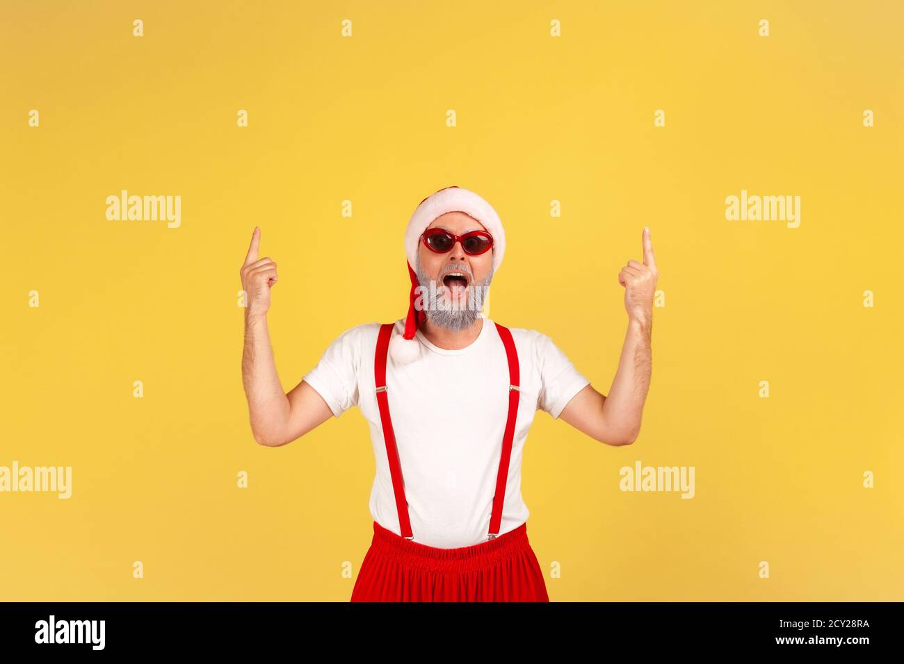 Extremely happy positive elderly man in santa claus hat and stylish sunglasses pointing fingers up at empty space for your advertising. Indoor studio Stock Photo