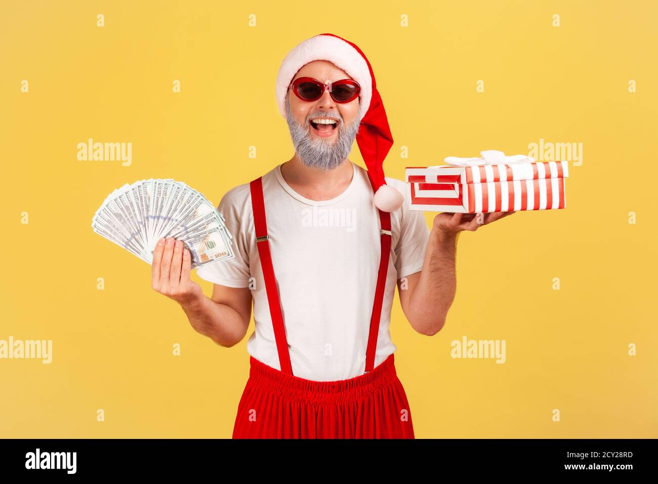 Cheerful positive elderly man in sunglasses and santa claus hat holding fan of dollars and gift box smiling at camera, preparing presents on holidays. Stock Photo