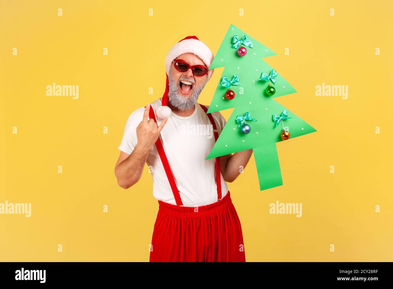 Extremely happy excited gray bearded man in santa claus hat holding paper christmas tree showing rock and roll gesture, having fun on winter holidays. Stock Photo