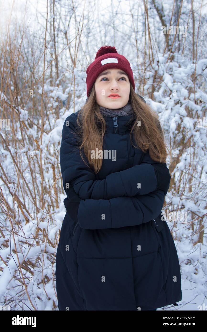 Beautiful girl freezing in winter forest. Portrait of serious cute attractive young teen girl with long blonde hair with green eyes on natural blurred Stock Photo