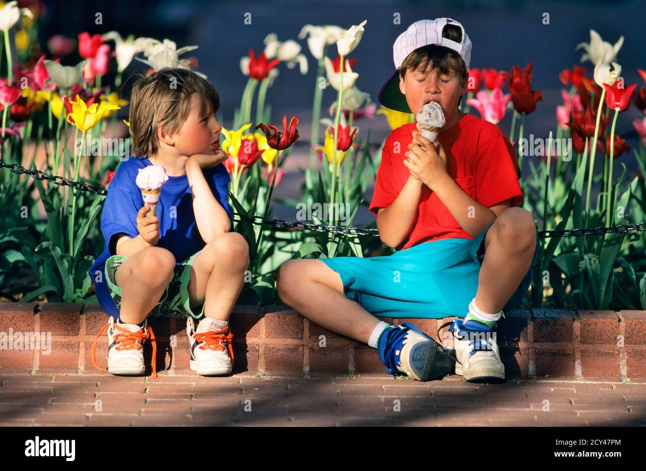 1990s TWO BOYS SITTING ON BRICK SIDEWALK BY TULIPS EATING ICE CREAM CONES YOUNGER BOY NOT EATING CONE STARING AT HIS BROTHER - kf29764 WAL004 HARS COPY SPACE FRIENDSHIP HALF-LENGTH PERSONS MALES SNEAKERS SIBLINGS LICKING STARING HAPPINESS HIS LEISURE CHOICE TULIPS NOT CONES SIBLING CONNECTION CONCEPTUAL NOURISHMENT ICE CREAM BACKWARD BASEBALL CAP GROWTH JUVENILES LICK RELAXATION SPRINGTIME TOGETHERNESS YOUNGER CAUCASIAN ETHNICITY FLAVOR OLD FASHIONED Stock Photo