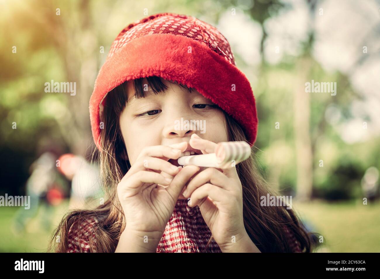 Happy little school girl playing party blow horn on her birthday in the garden in summer. Childhood lifestyles concept. Stock Photo