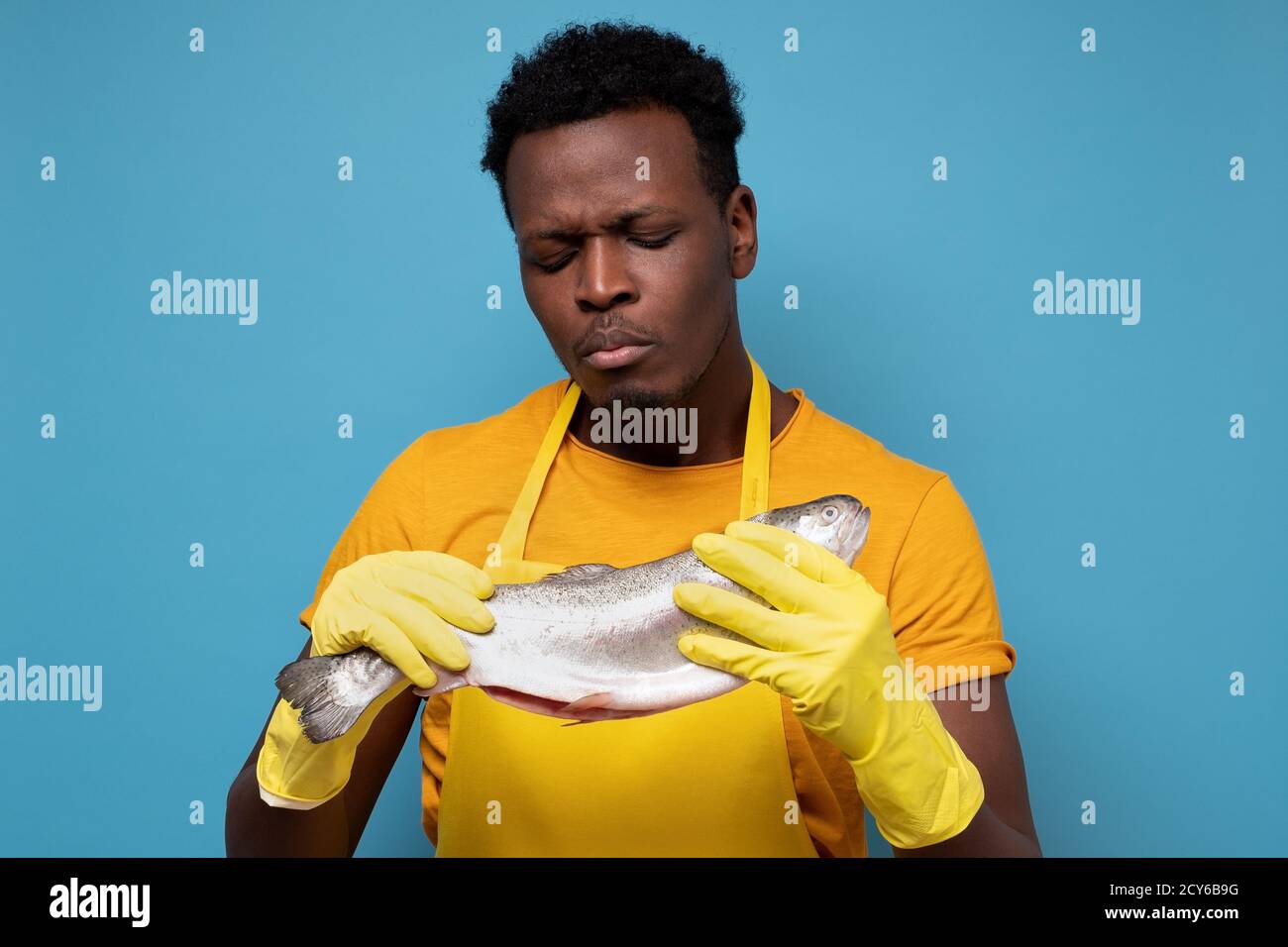 African young man in yellow apron and gloves examining smelly salmon fish Stock Photo