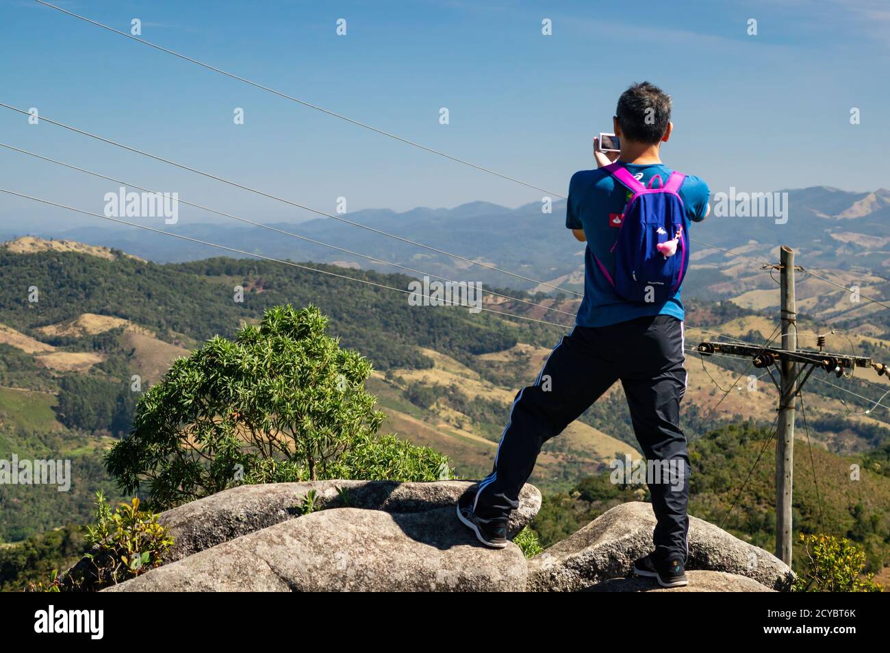 A man standing on a big rock taking pictures of the mountainous Serra do Mar (Sea Ridge) mountainous landscape at the top of Pedra da Macela landmark. Stock Photo