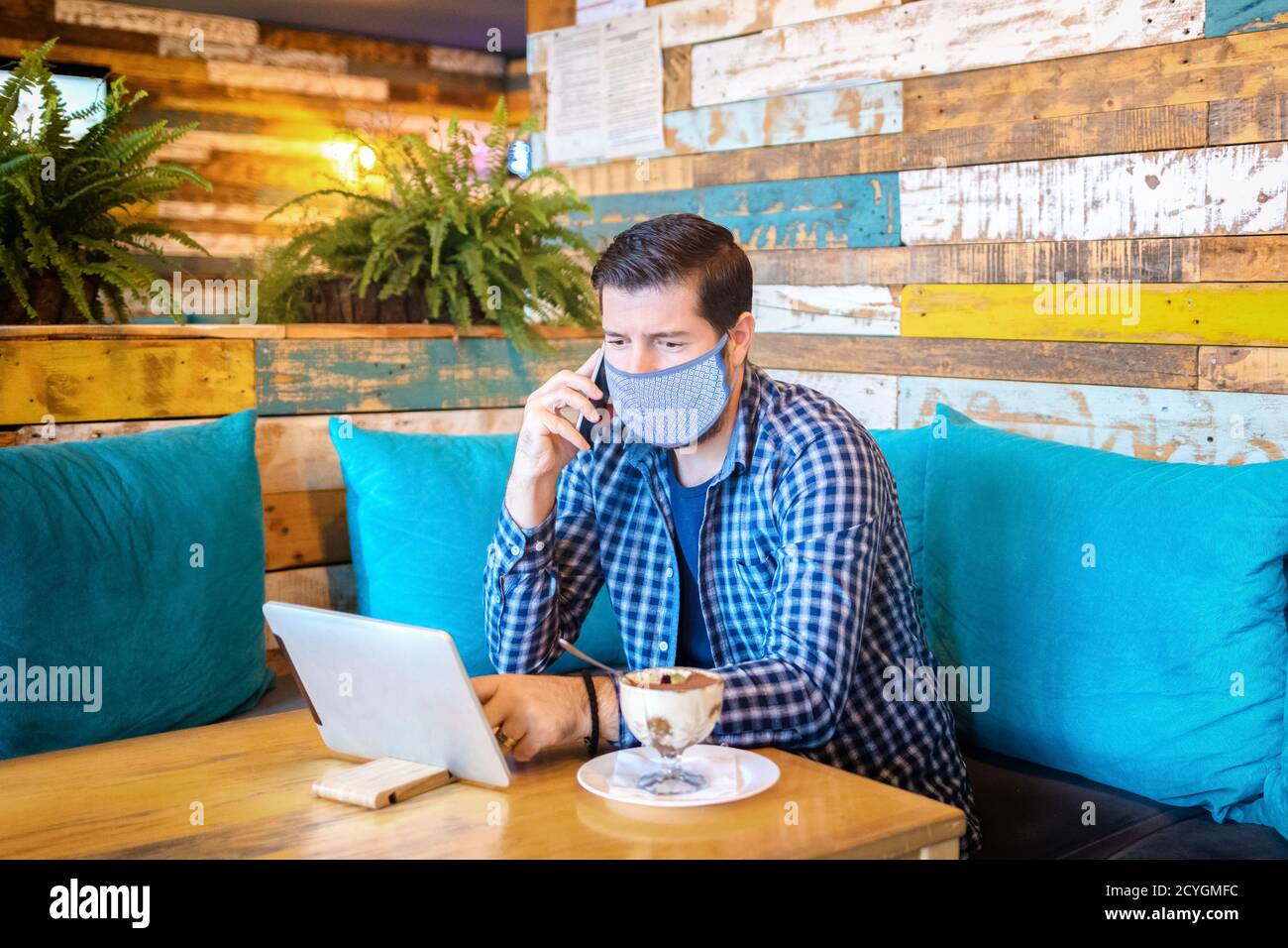 Relaxed entrepreneur with protective face mask remote working at digital tablet while talking on mobile phone in coffee shop Stock Photo