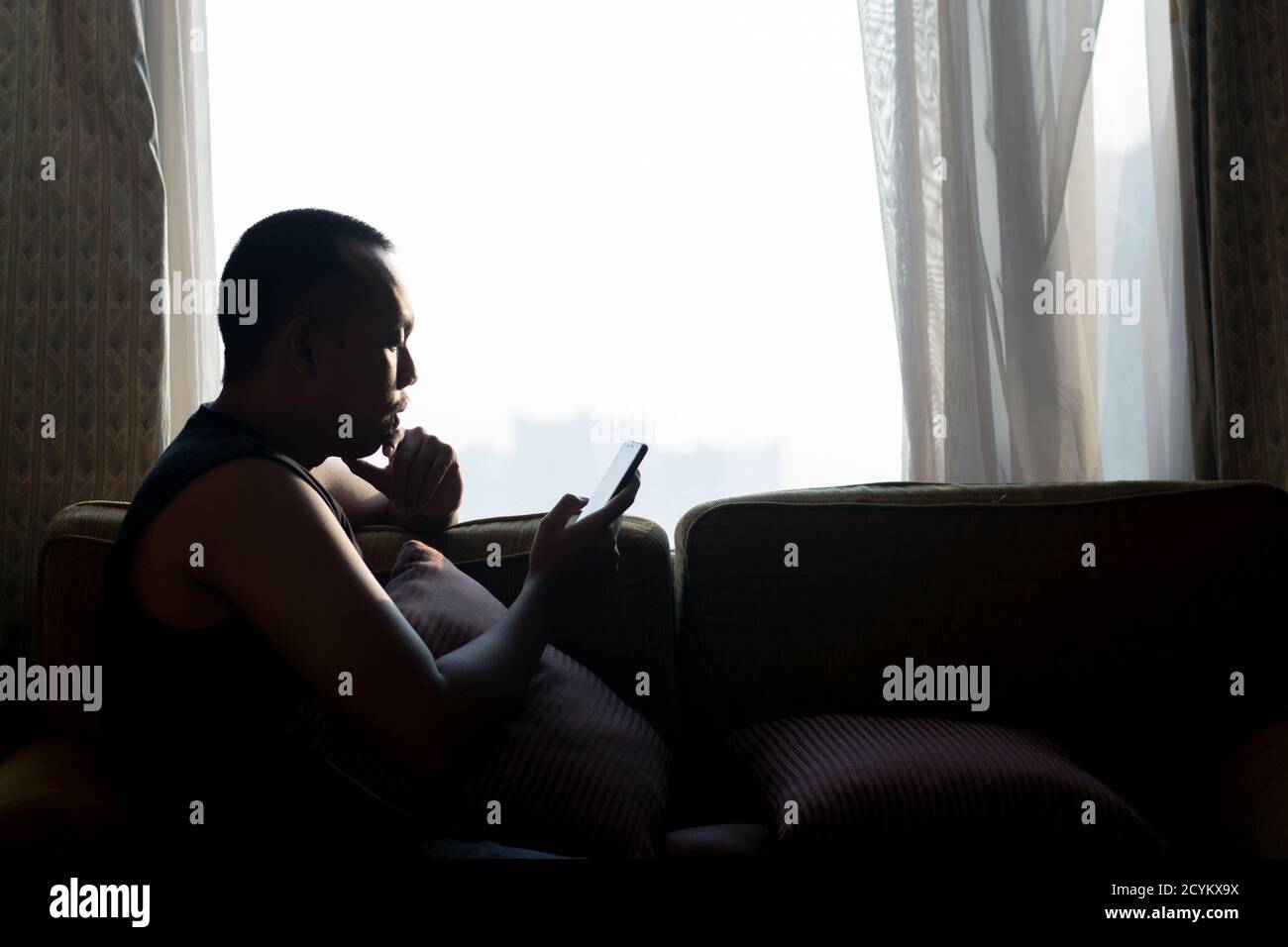 Asian man in armless clothes sitting in room near clear glass window, eyes looking at smart phone and using mobile phone in hand holding Stock Photo