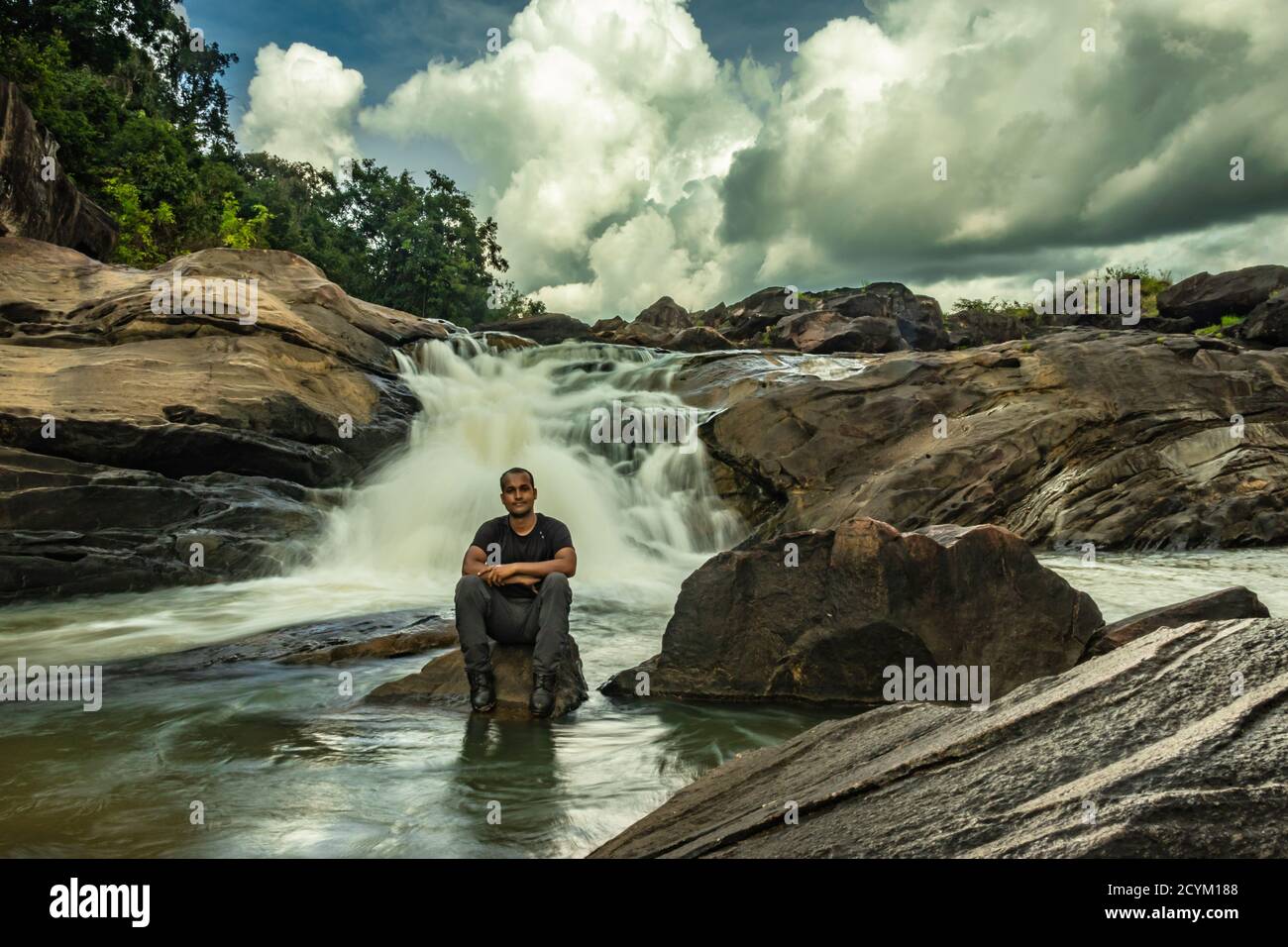 man sitting at rock watching the beautiful waterfall stream at evening image is taken at remote western ghat forests karnataka india. it is showing th Stock Photo