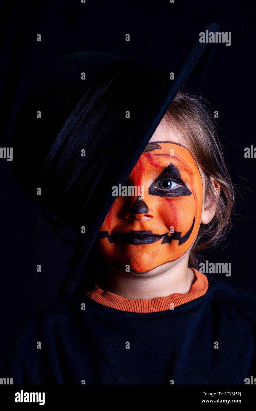 girl's face with pumpkin makeup in black hat on black background, isolated, Halloween celebration on self-isolation Stock Photo