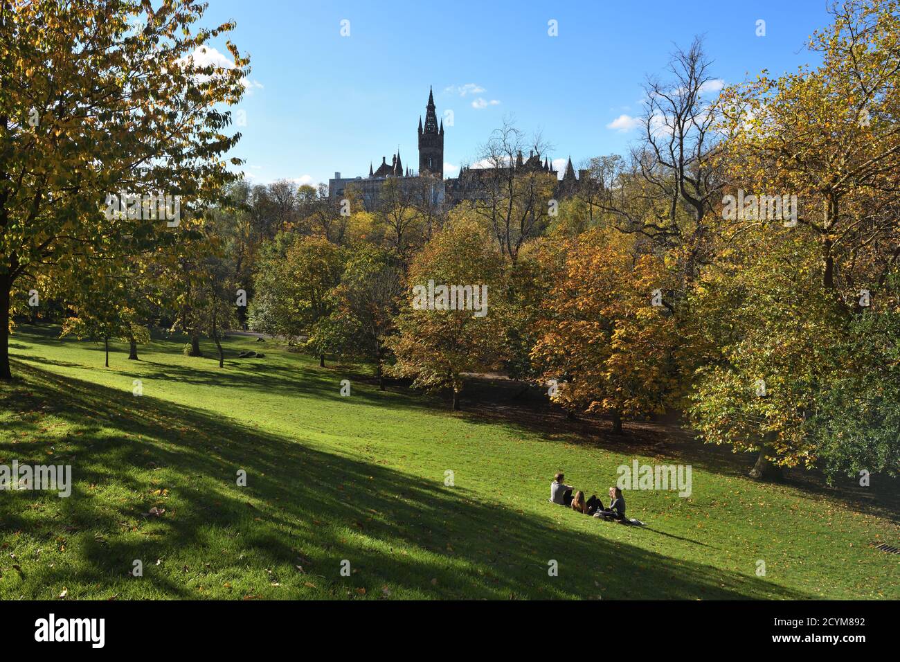 Three female student relaxing in the Autumn sunshine in Kelvingrove Park with Glasgow University building, Scotland, UK Europe in the background Stock Photo