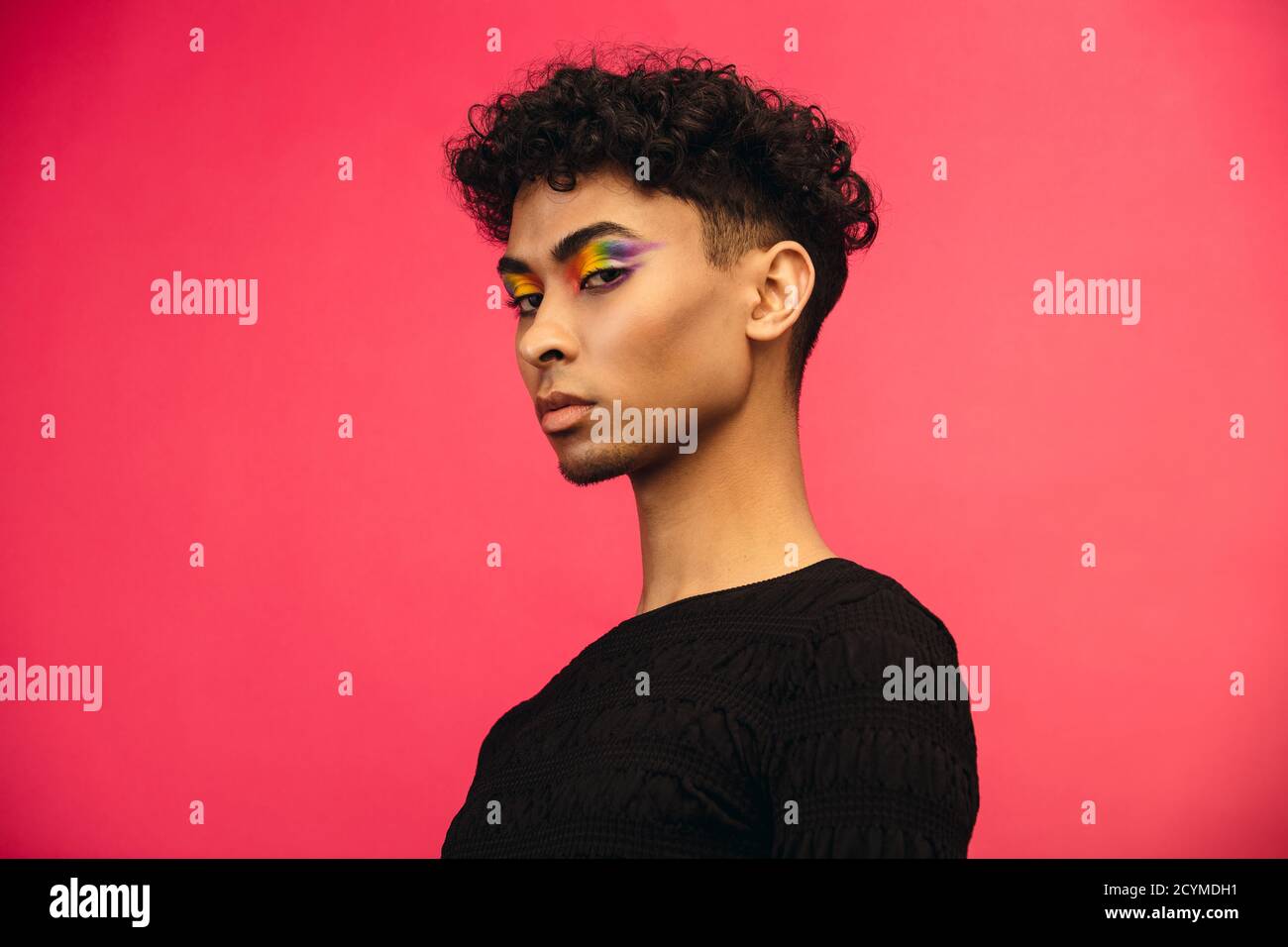 Handsome man in black t-shirt staring at camera. Androgynous man wearing black outfit and rainbow colored eye shadow against red background. Stock Photo