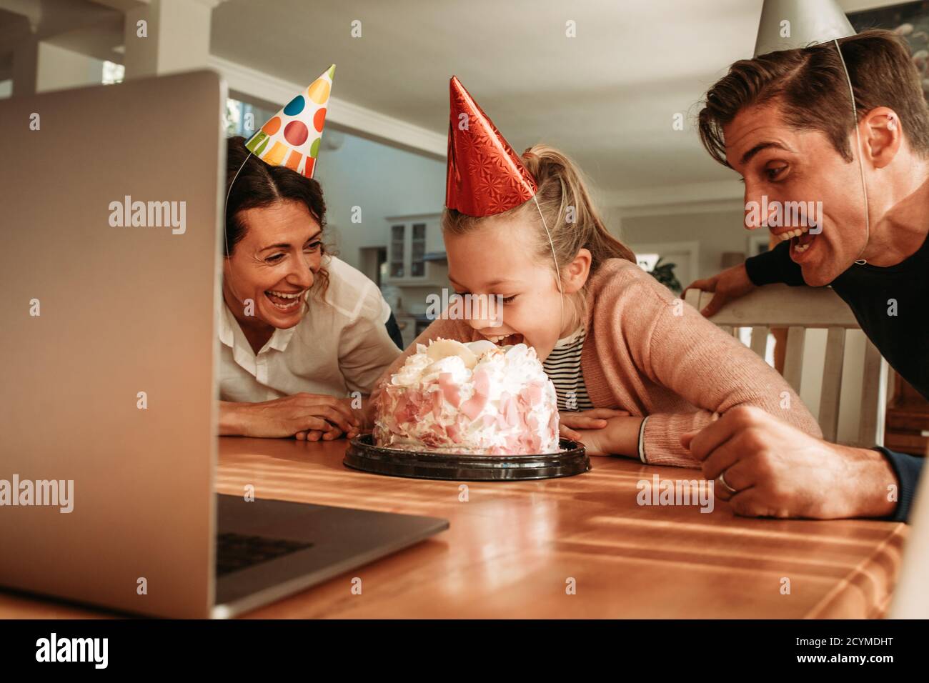Girl cutting her birthday cake with her mouth with her parents smiling by. Family celebrating girl's at home during covid-19 quarantine. Stock Photo