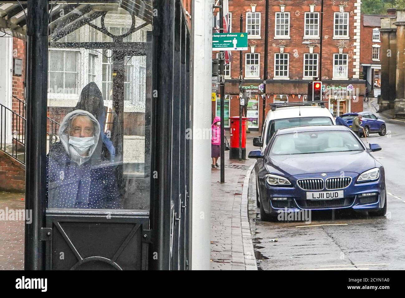 Bewdley, UK. 2nd October, 2020. UK weather: persistent rain and breezy winds make for a miserable shopping experience on an already quiet high street. A lady looks truly unhappy while wearing a mask using the cover of the bus shelter to protect her from the wind and rain as she waits for her transport home. Credit: Lee Hudson/Alamy Live News Stock Photo