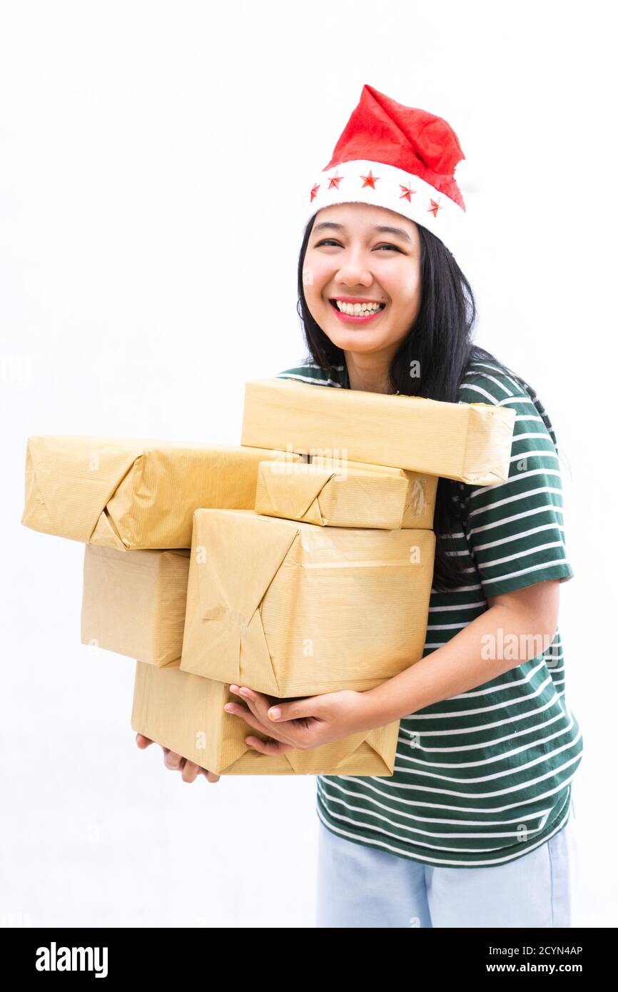 portrait of a young Asian woman wearing a santa hat and wearing casual clothes lifting a pile of gifts with a white isolated background Stock Photo