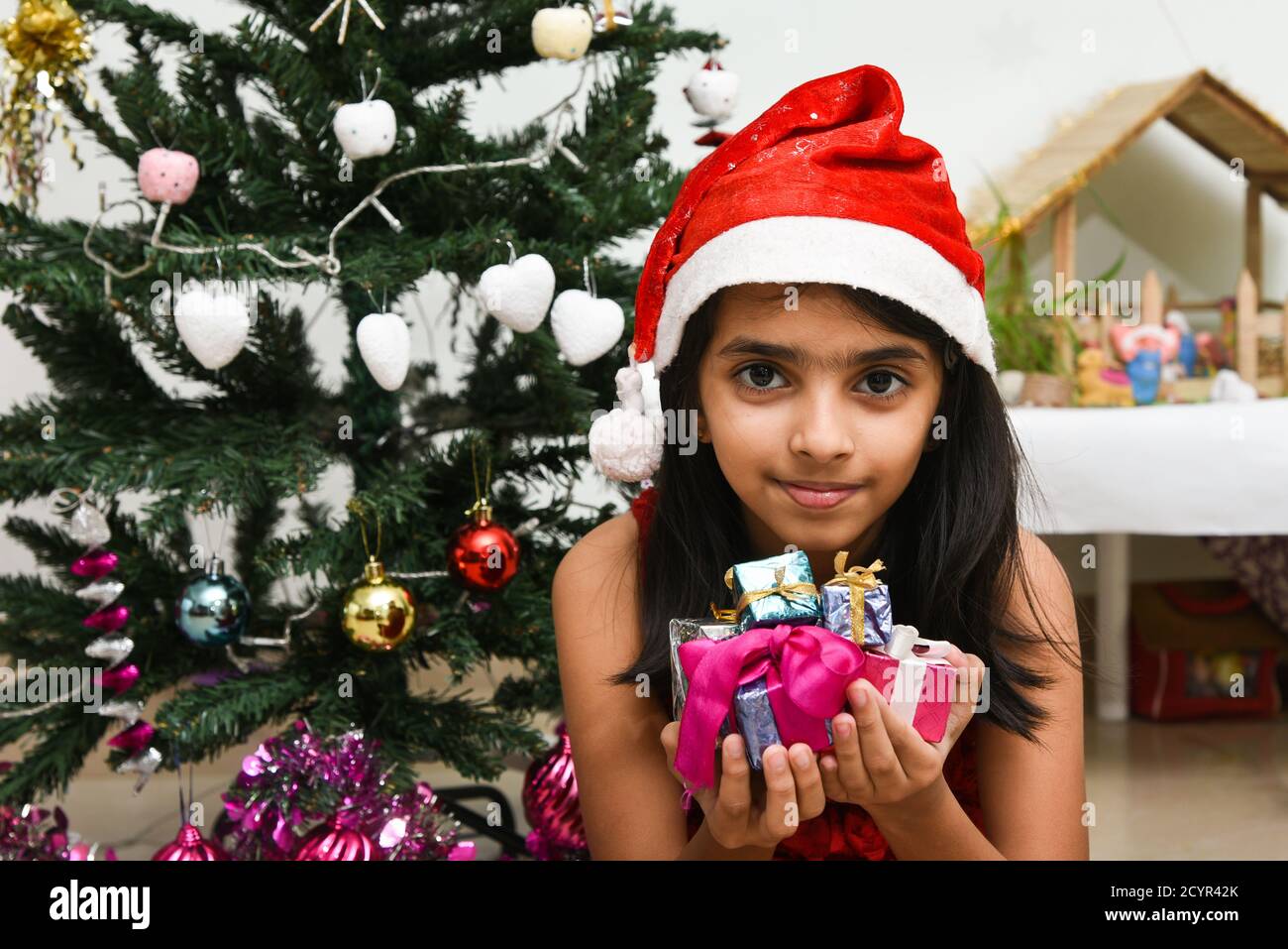 cute little Indian girl celebrating Christmas in India many present Indian Christmas tree. Happy child wearing Santa helper red hat with lot of x-mas Stock Photo