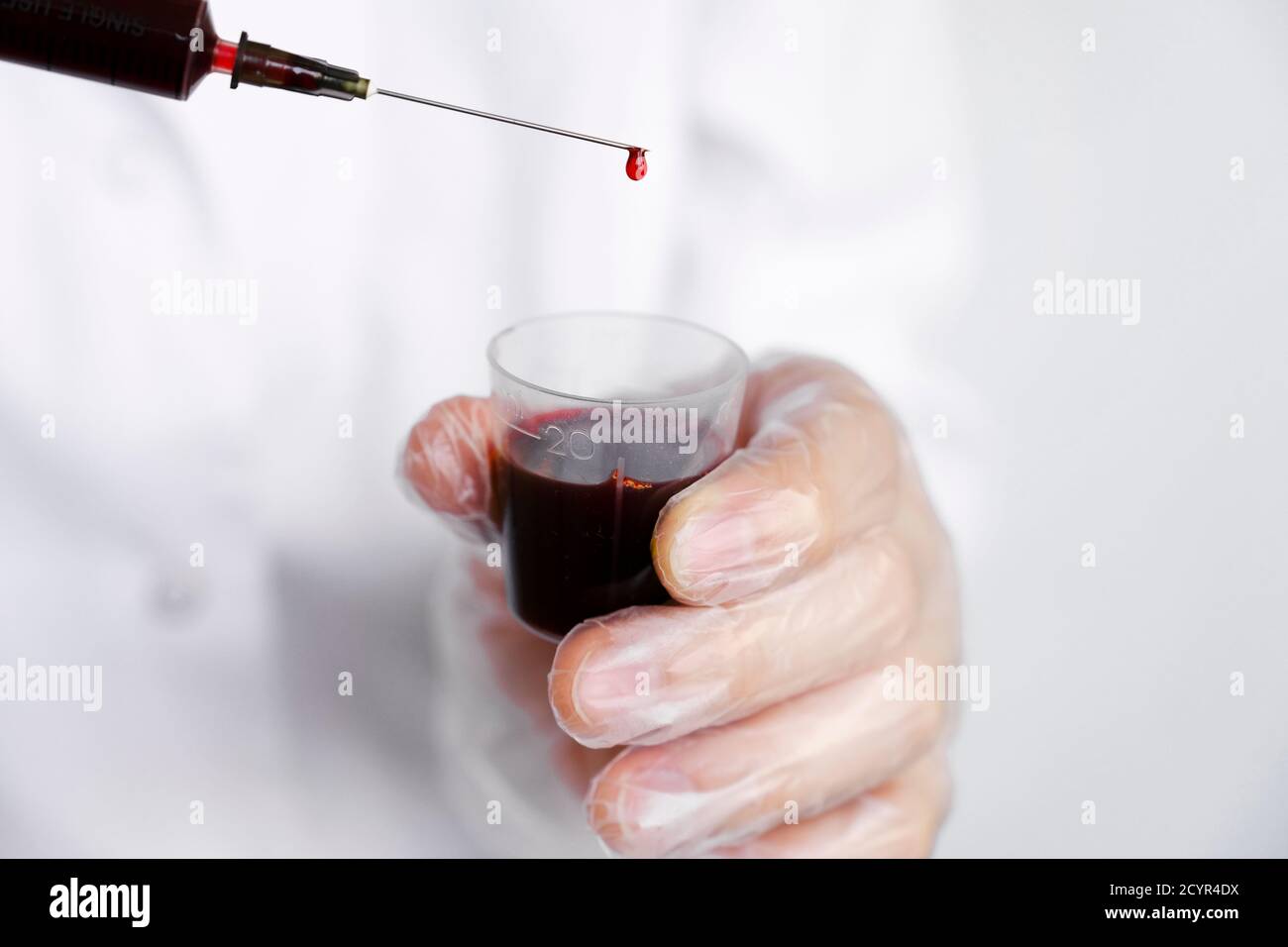 Doctor or lab scientist in personal protective equipment holding a syringe full of blood, with focus on single drop dripping out of the needle tip Stock Photo