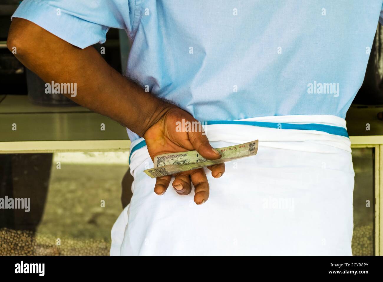 Man wearing lungi at a Main Road shop counter holding 100 rupee note, in this busy town in Wayanad district; Kalpetta, Wayanad, Kerala, India Stock Photo