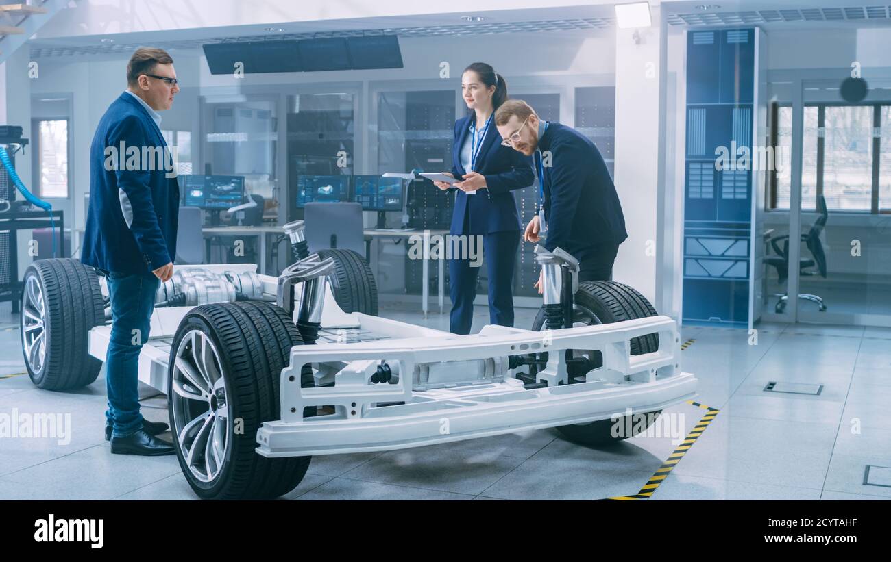 Automotive Design Engineers Talking while Working on Electric Car Chassis Prototype. In Innovation Laboratory Facility Concept Vehicle Frame Includes Stock Photo