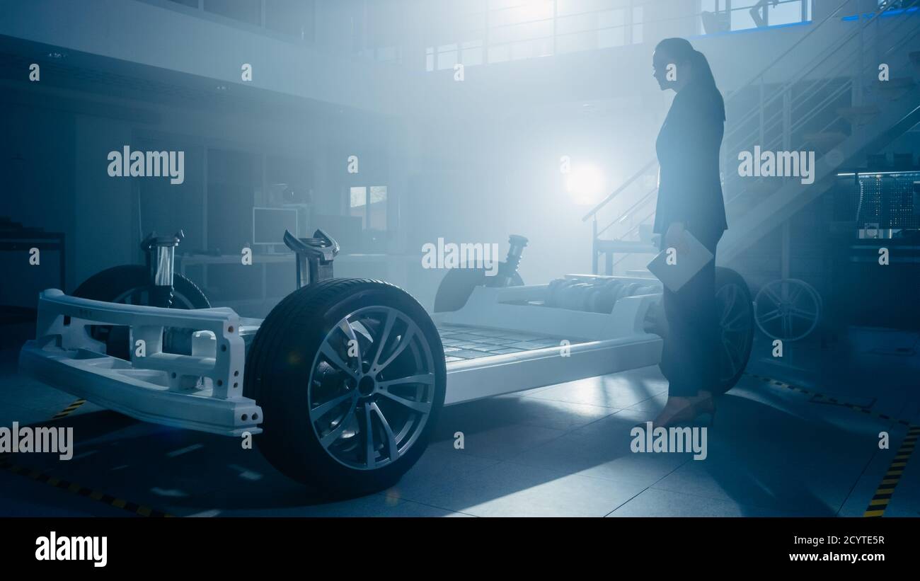 Silhouette of a Female Automobile Design Engineer in Automotive Innovation Facility Working on Electric Car Platform Chassis Prototype that Includes Stock Photo
