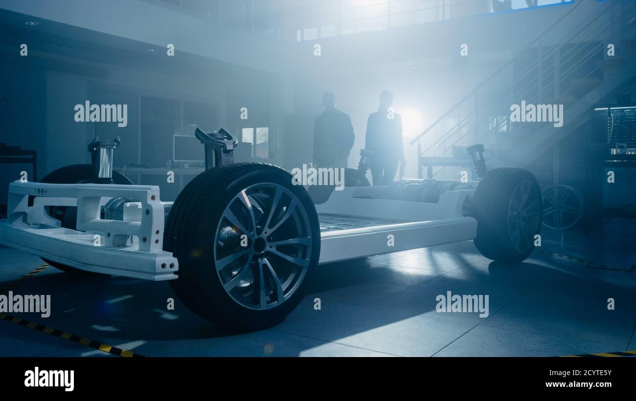Silhouettes of Two Automobile Design Engineers In Automotive Innovation Facility. They are Working on Electric Car Platform Chassis Prototype that Stock Photo