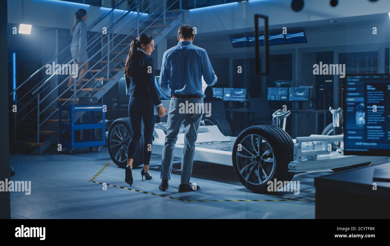 Auto Industry Design Facility: Male Chief Engineer Shows Car Prototype to Female Car Designer. Electric Vehicle Platform Chassis Concept Has Wheels Stock Photo