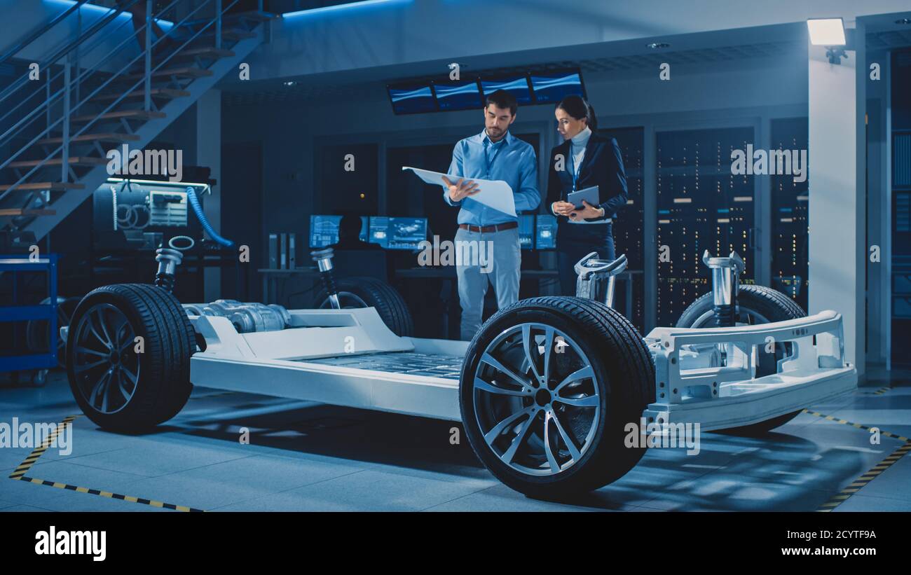 Auto Industry Design Facility: Male Chief Engineer Shows Car Blueprints Female Software Design and Integration Engineer. Electric Vehicle Platform Stock Photo