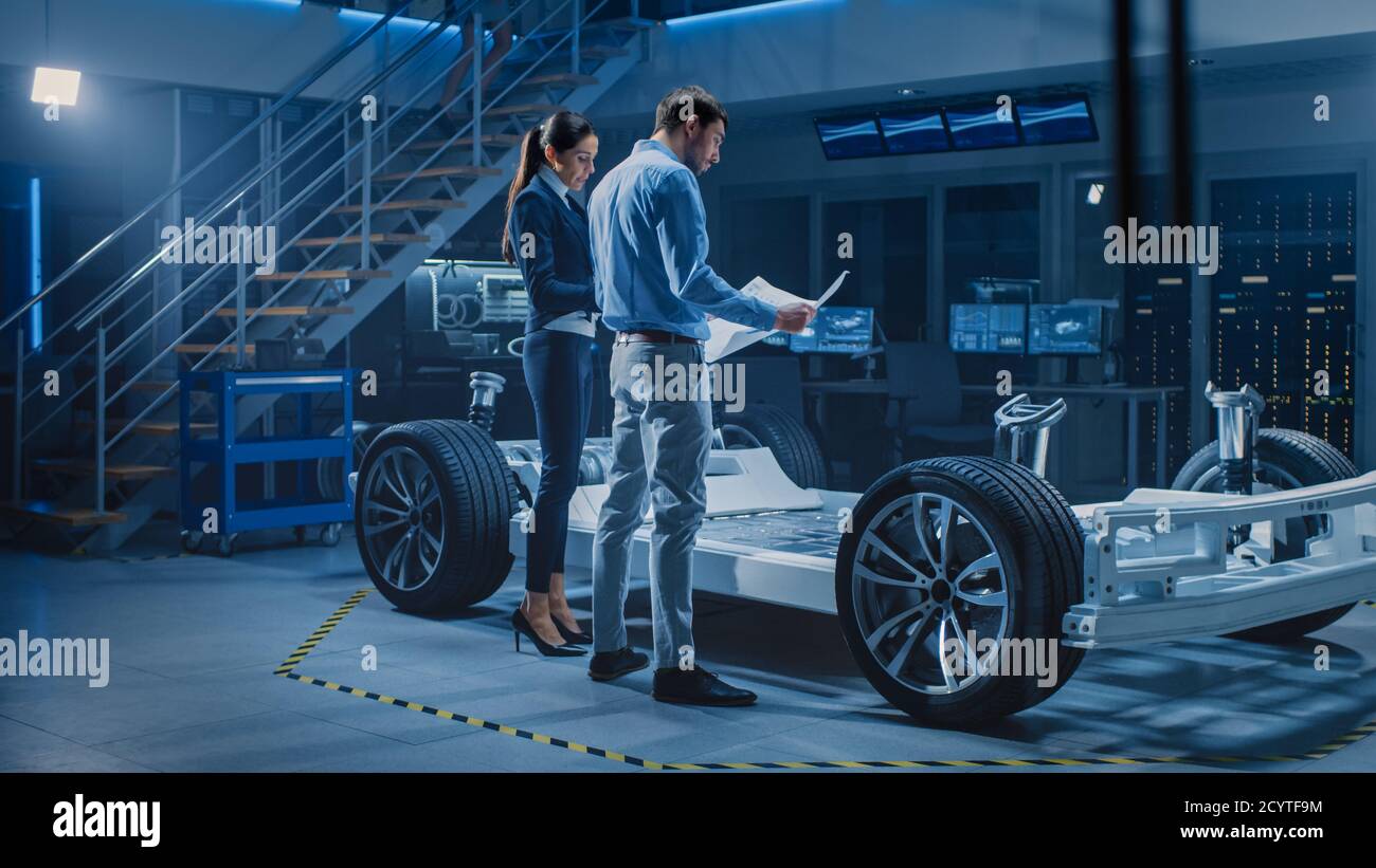 Auto Industry Design Facility: Male Chief Engineer Shows Car Blueprints Female Software Design and Integration Engineer. Electric Vehicle Platform Stock Photo