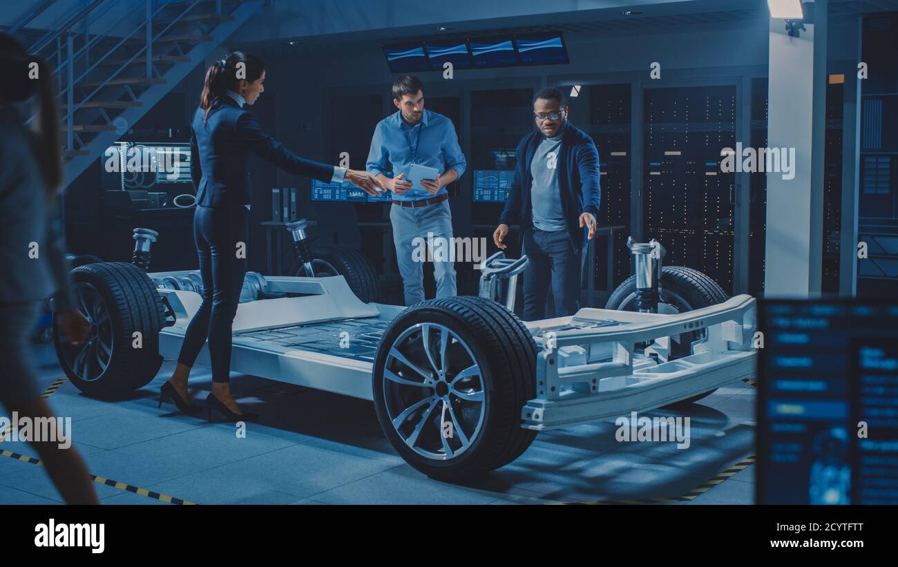 Female Project Manager Talks about Electric Car Prototype with Her Team of Engineers, Showing Chassis, Explaining Sustainability and Ecological Stock Photo