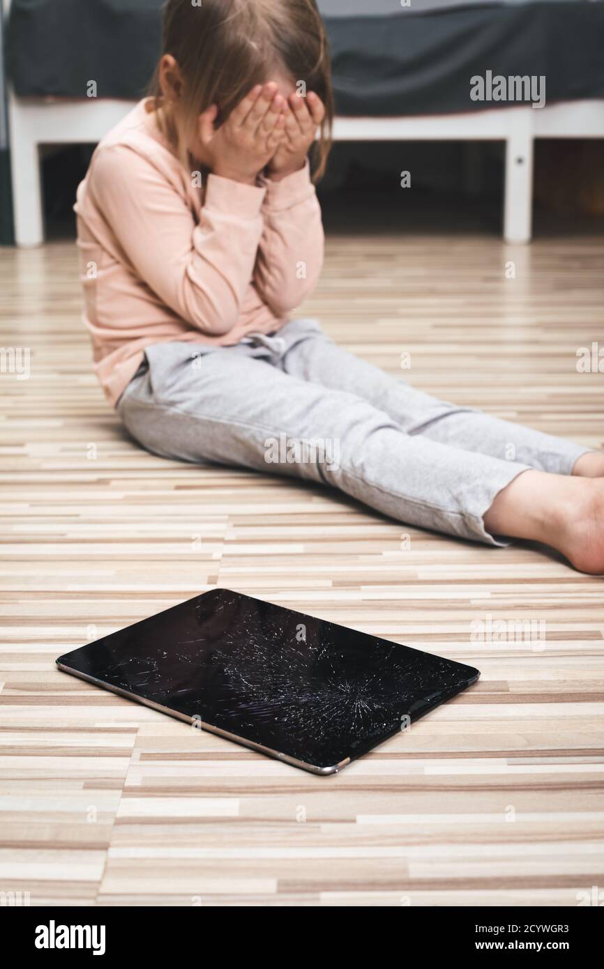 Little gird accidentally broke a tablet computer. Focus on cracked touch screen of a tablet. Upset child is crying in background afraid of punishment Stock Photo