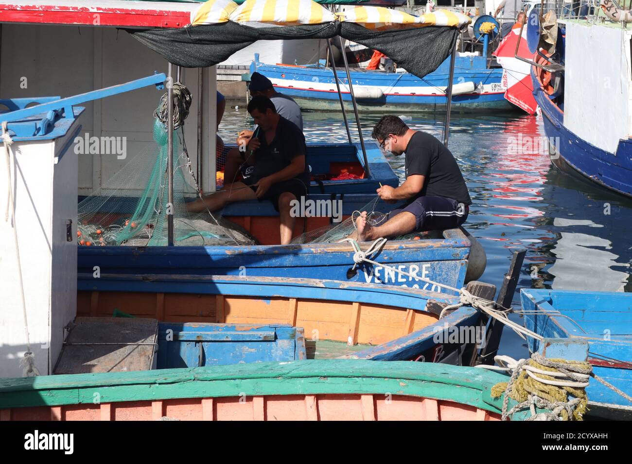 Fishermen mending their fishing nets on the decks of their boats in the harbor in Trapani Old Town Sicily Stock Photo