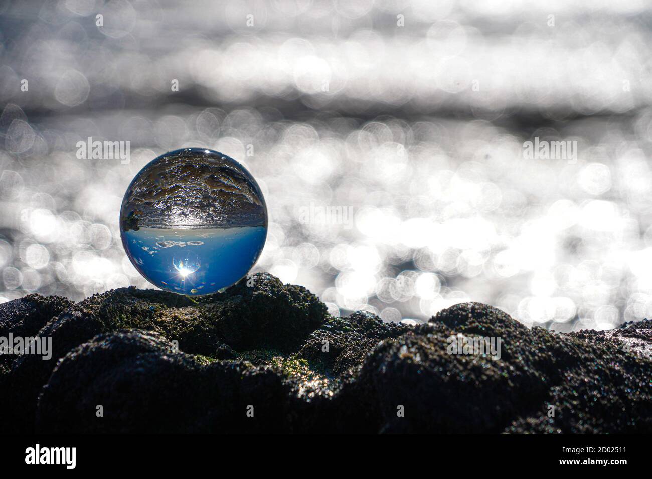Photography Crystal Photo Ball showing upside down reflection of sky beach and waves balanced on rocks on sandy beach. Stock Photo
