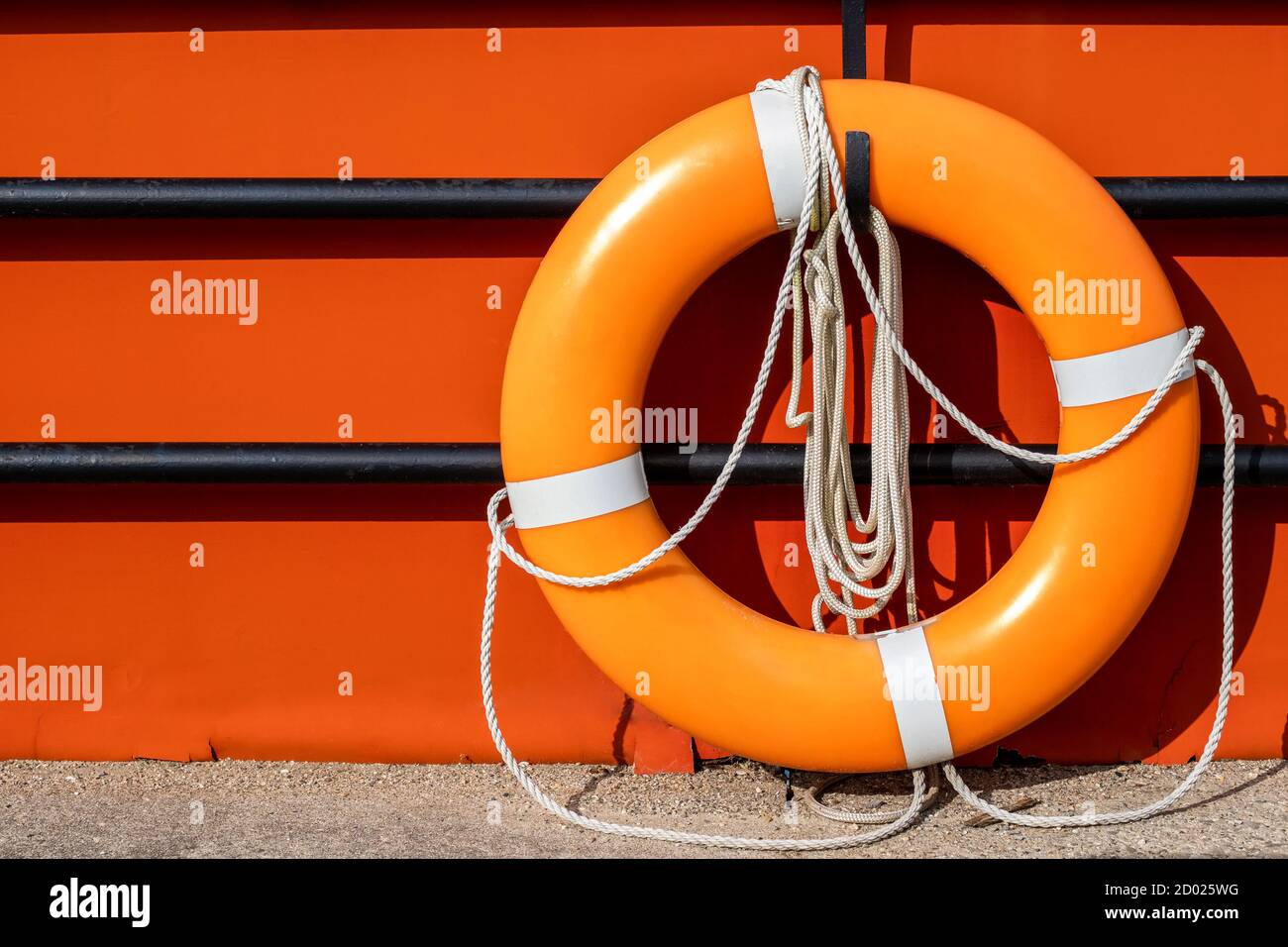 An orange life ring hangs on a hook on a black fence in front of an orange background surface. The life preserver has white ropes attached. Stock Photo