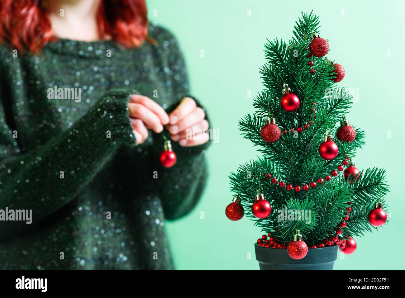 Little Christmas tree in a pot decorated with red Christmas baubles. Young girl in a green and cozy sweater decorating the Christmas tree. Stock Photo