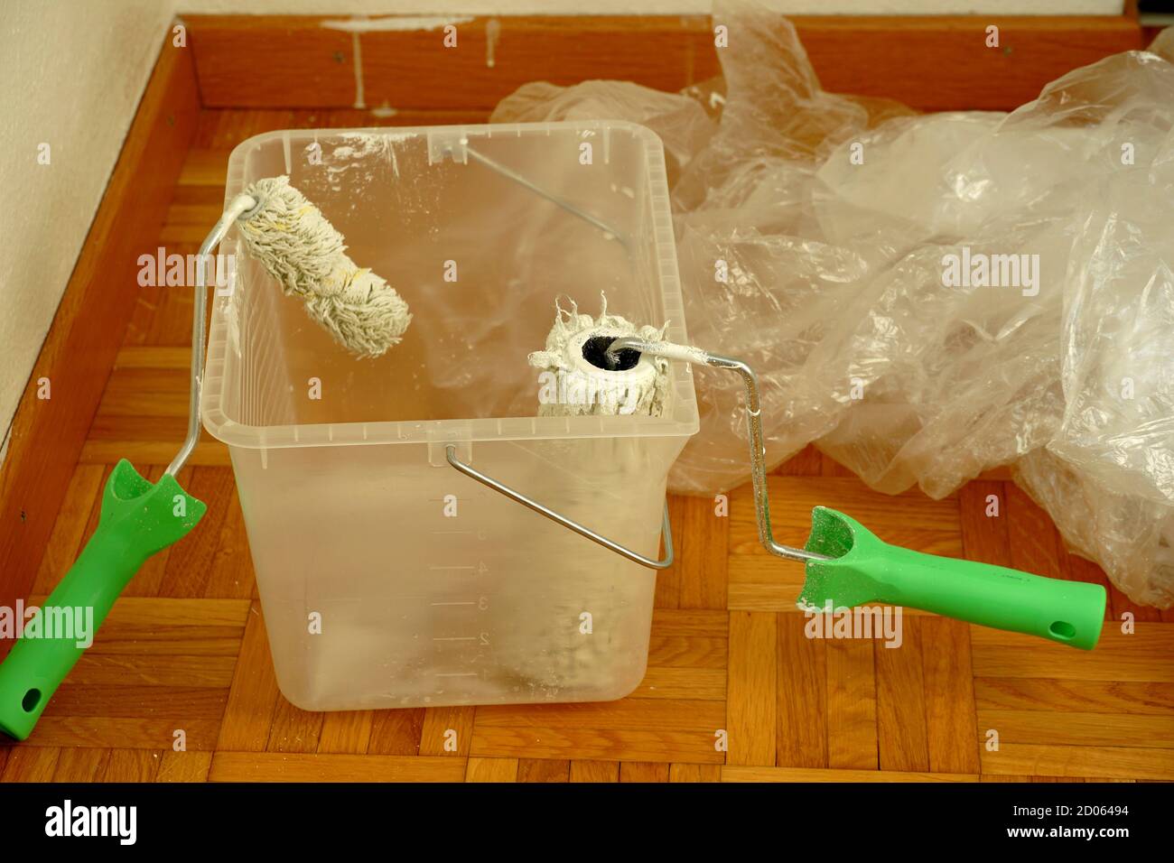 Two paint rollers in a container on the floor. The paint is dripping off the rollers. In background there is a plastic foil to protect the furniture. Stock Photo