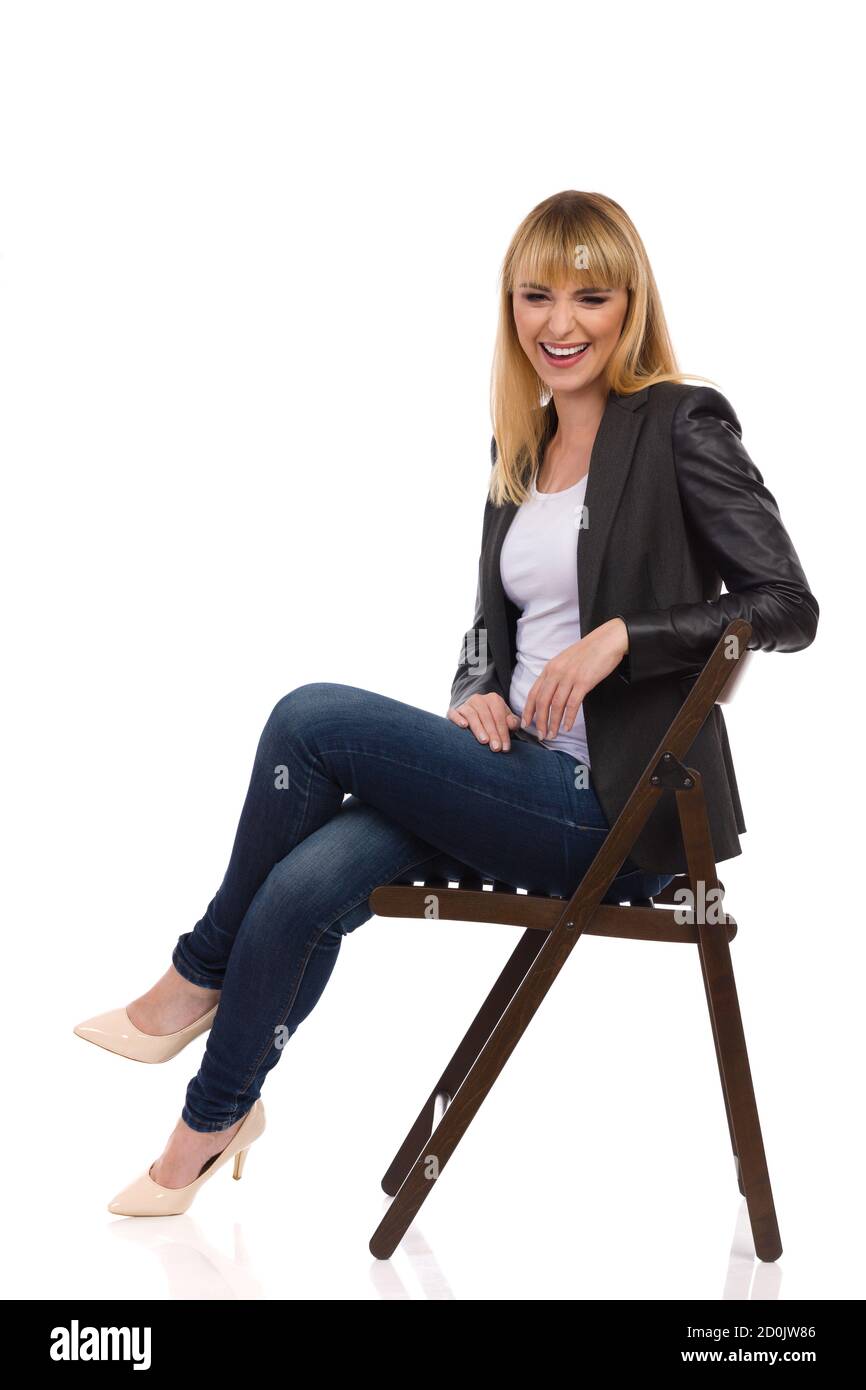 Laughing young woman is sitting on a chair and looking at camera. Side view. Full length studio shot isolated on white. Stock Photo