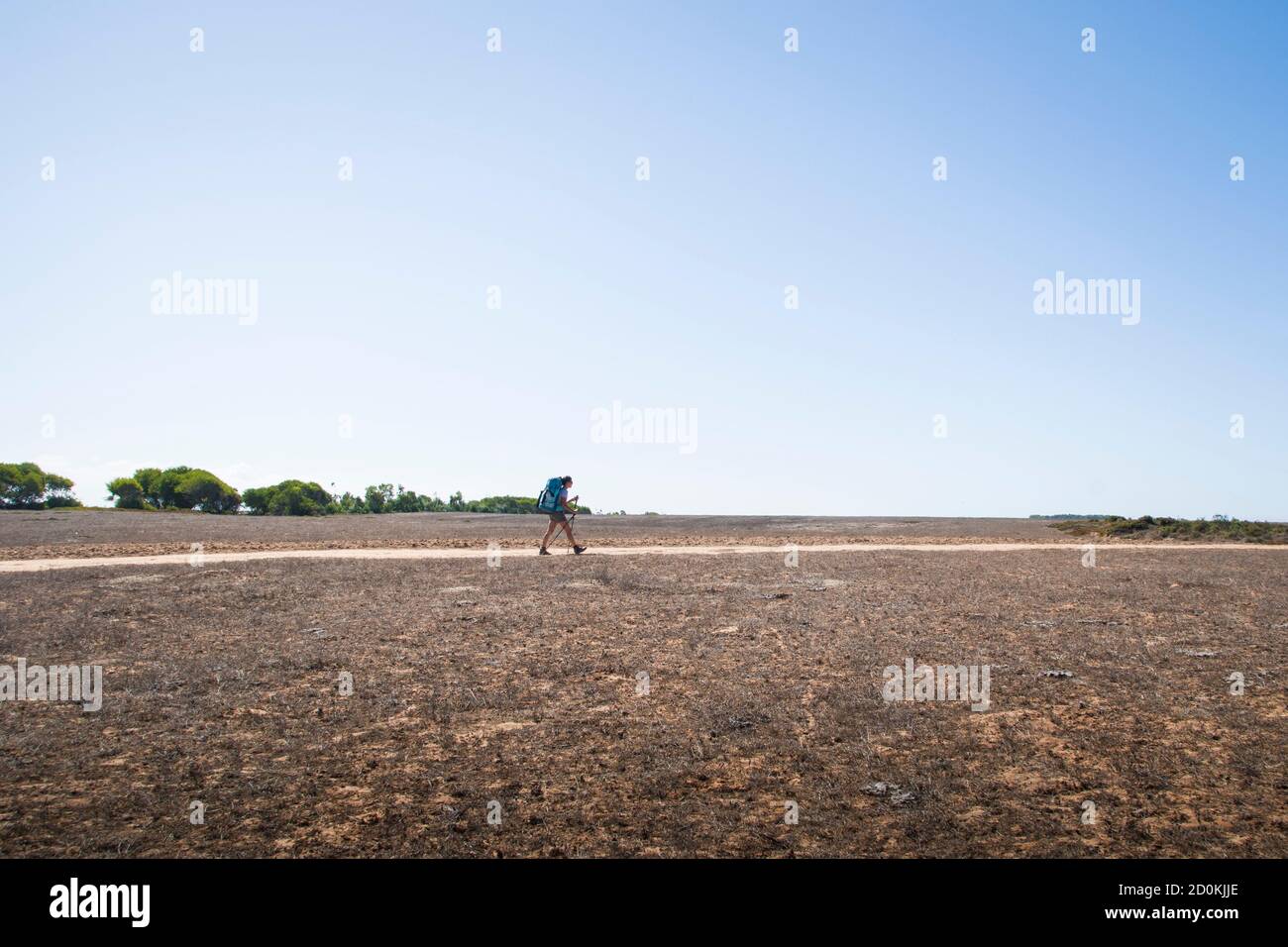 Hiker walks with backpack on dusty road in a vast dry landscape during hard daylight Stock Photo