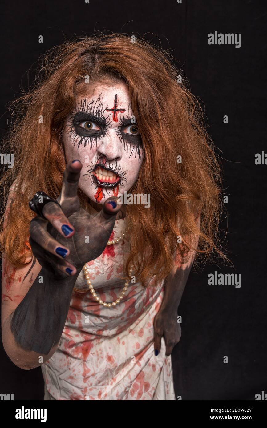 A young woman with red hair and professional make-up to look creepy, wearing bloody white clothes. On a black background Stock Photo